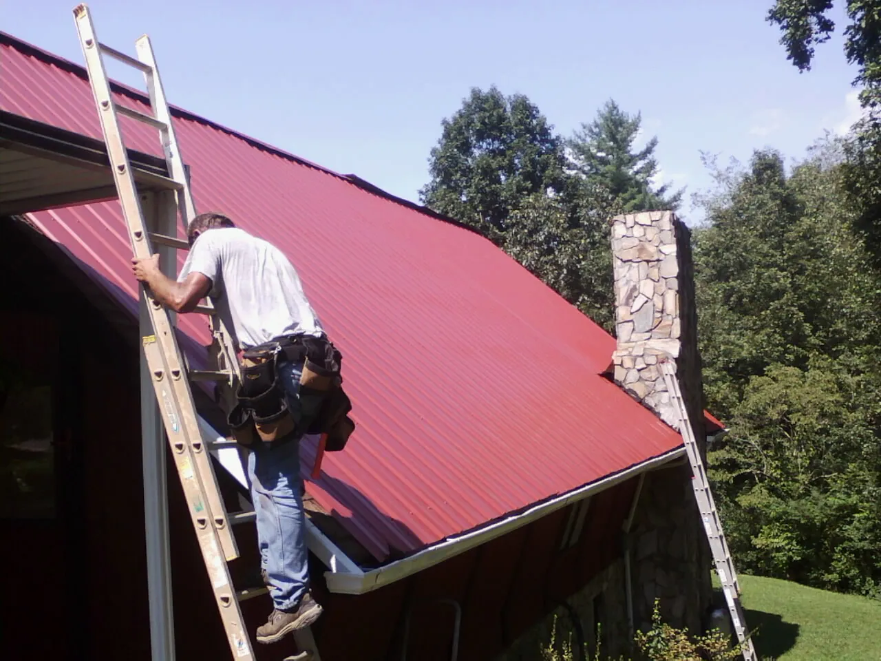 Roofer on a ladder beside a red metal roof and stone chimney — Soffit & Fascia Repair by Roofing Brothers in Sanderson